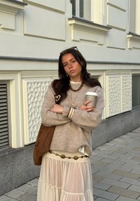 Pulover bej tricotat, fustă crem cu design stratificat și curea decorativă, geantă tote maro, ceașcă gri, fundal urban cu perete texturat.