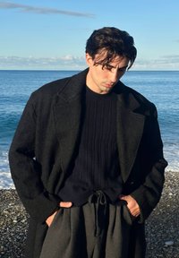 Manteau noir texturé par-dessus un pull en maille torsadée bleu marine, associé à un pantalon gris à cordon de serrage. Le modèle est debout près de l'eau, les mains dans les poches.