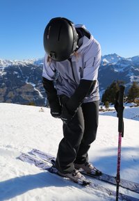L'équipement de ski comprend un casque noir, une veste grise et un pantalon noir. Les skis sont violets avec des fixations. Un bâton de ski présente un accent rose. Arrière-plan de montagne enneigée.