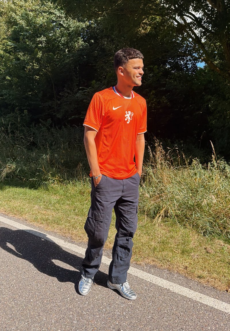 Jeune homme portant un maillot de sport orange et un pantalon cargo foncé, souriant, debout au bord d'une route ensoleillée avec des arbres et de l'herbe derrière lui.