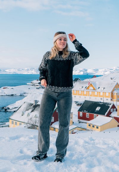 Suéter de lana negro con un yugo estampado en blanco, combinado con jeans grises y botas negras, en un paisaje nevado con casas coloridas.