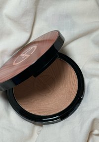 Poudre bronzante compacte avec surface texturée et logo en relief, boîtier rond noir et or rosé, placé sur un tissu blanc doux.