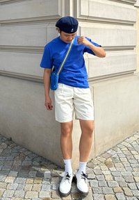 T-shirt di cotone blu, pantaloni corti bianchi, scarpe in bianco e nero, calzini bianchi. Piccola borsa blu con tracolla gialla. Muro di pietra e ciottoli sotto.