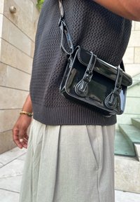 Μαύρη παπουτσοθήκη από γυαλιστερό δέρμα με διπλές αγκράφες, συμπαγές ορθογώνιο σχήμα και ρυθμιζόμενο λουρί, φορεμένη πάνω από μια υφασμάτινη ζακέτα.