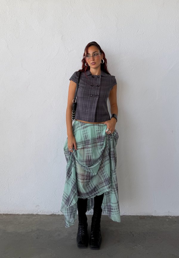 Une femme porte un haut à carreaux gris, une jupe fluide à carreaux vert menthe, des bottes noires, des lunettes et un sac à bandoulière, devant un mur blanc.