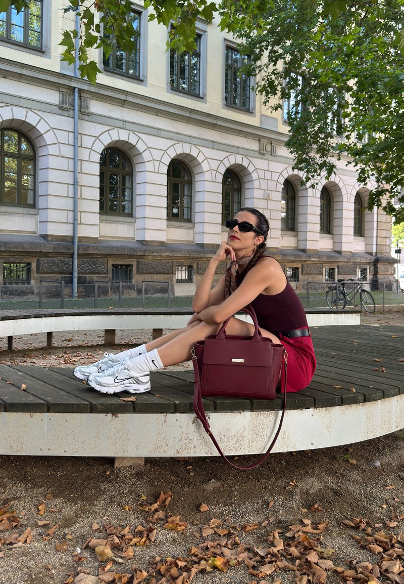 Jovem mulher com óculos de sol sentada num banco circular perto de um edifício, com uma mala bordeaux, ténis e folhas caídas no chão.
