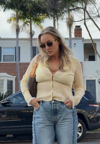 Γυναίκα με ξανθά μαλλιά, φοράει γυαλιά ηλίου, ένα κρεμ πουκάμισο με κουμπιά και μπλε τζιν, στέκεται με τα χέρια στις τσέπες δίπλα σε ένα μαύρο SUV και κατοικίες.