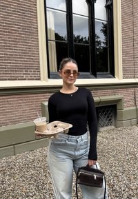 Wiederverwendbarer Kaffeebecher mit einem klaren Deckel, der in einer Hand gehalten wird. Die andere Hand hält ein beige Papptablett. Der Hintergrund zeigt eine strukturierte Ziegelwand.