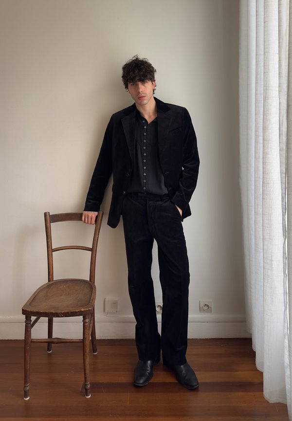 Jonge man in een zwart pak en overhemd leunt op een houten stoel, staande op een houten vloer naast een wit gordijn en wandcontactdozen.