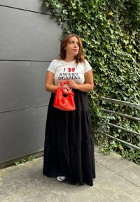Hvid t-shirt med sort tekst, der står "I SWEET DRAMAS," parret med en lang sort nederdel. Holder en lille rød taske. Grøn ivybakgrund.