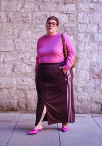 Μωβ πουλόβερ, καφέ αθλητική φούστα με ροζ πλαϊνές ρίγες, ροζ μύτες, κοντά μαλλιά, στέκεται μπροστά σε έναν τοίχο από πέτρα.