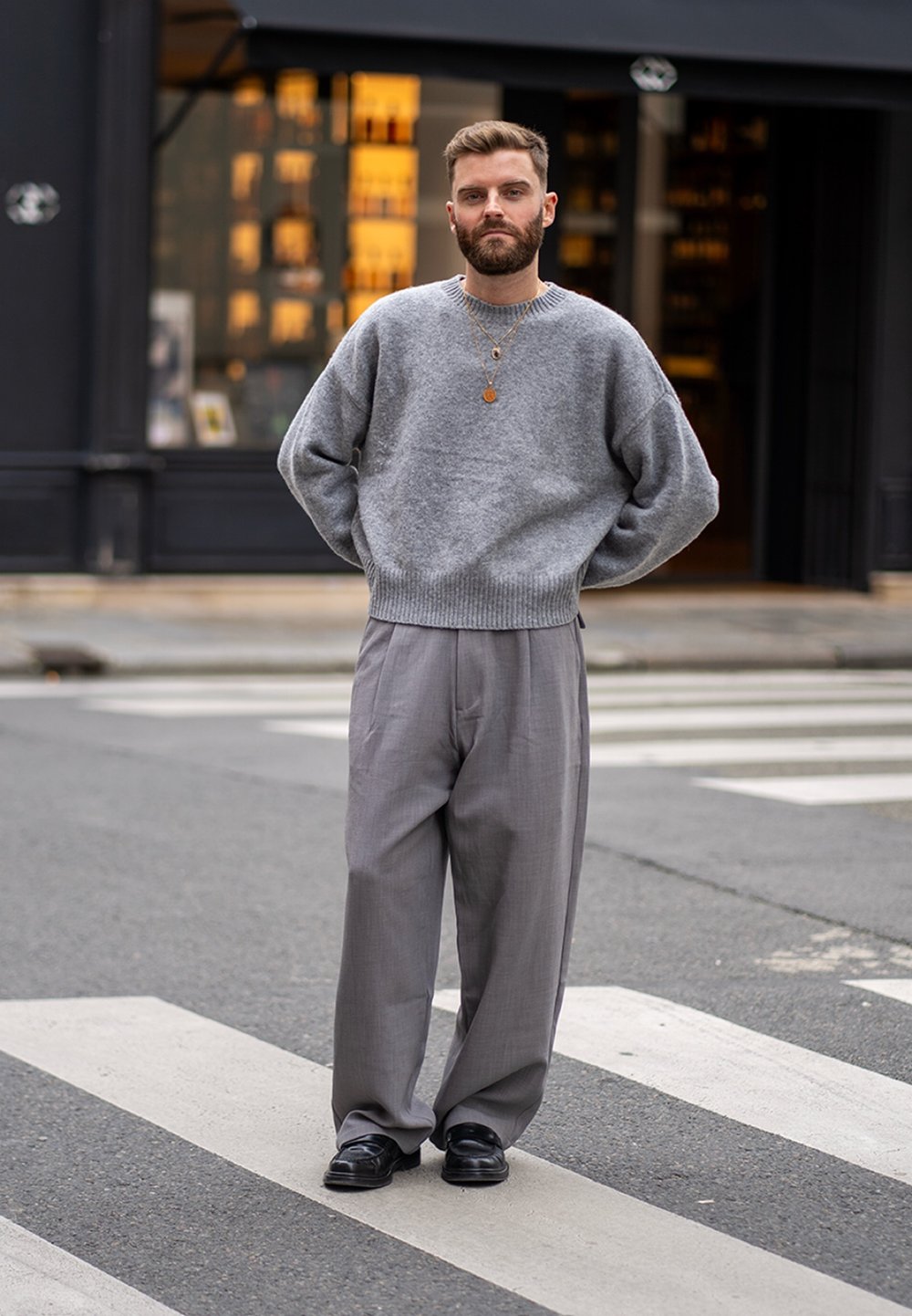 Pulover gri tricotat, pantaloni gri largi, pantofi negri, colier cu pandantiv, stând pe o trecere de pietoni cu un magazin în fundal.