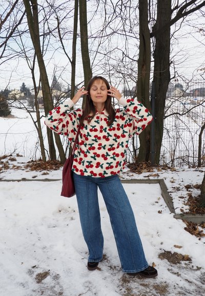 Mujer de pie sobre suelo nevado, vistiendo un suéter de forro polar blanco con patrones de cerezas rojas, vaqueros azules y un bolso rojo al hombro, con los ojos cerrados.