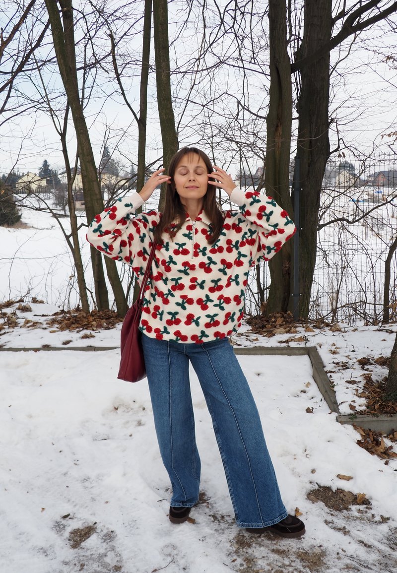 Femme debout sur un sol enneigé, portant un pull polaire blanc avec des motifs de cerises rouges, un jean bleu et un sac à bandoulière rouge, les yeux fermés.