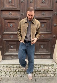 Veste en daim marron avec fermeture éclair, pull en maille grise, jean bleu et baskets grises. Posé devant une porte en bois ornée d'éléments décoratifs.