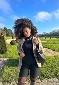 Beige blazer over en sort turtleneck, parret med sorte shorts. Tilbehør omfatter en perlekæde, øreringe og en sort håndtaske. Grøn landskabsbaggrund.