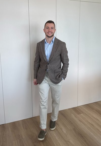 Joven sonriente, vestido con blazer marrón, camisa azul, pantalones blancos y zapatos verdes, de pie sobre suelo de madera frente a una pared blanca.