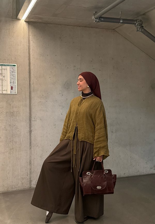 Donna con hijab marrone, cardigan senape e pantaloni larghi marroni, che tiene una borsa di pelle scura, in piedi contro un muro di cemento all’interno.