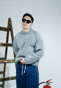 Pelēks un balts svītrains adīts džemperis ar apaļu kakla izgriezumu un ribotām aprocēm, kopā ar tumši ziliem sporta biksēm ar sānu svītrām.