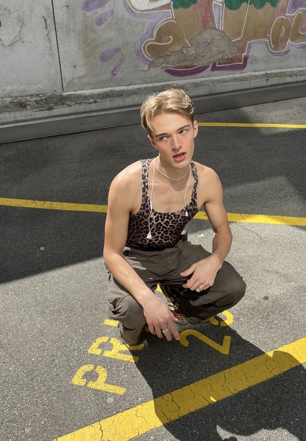 Junge Person mit blondem Haar, die ein Tanktop mit Leopardenmuster und Cargohosen trägt, hockt auf Asphalt nahe Graffiti und gelben Parkplatzmarkierungen.
