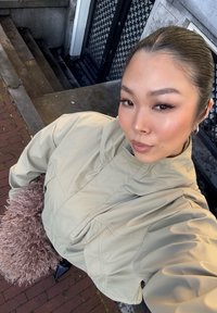 Jeune femme aux cheveux plaqués en arrière, portant une veste beige et tenant un sac brun duveteux, debout sur un trottoir en briques près d'escaliers en pierre.