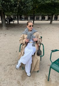 Beige Trenchcoat, grauer Schal, weißes Oberteil und weite Hose. Graue Sneaker. Sitzt auf einem grünen Stuhl in einem sandigen Park mit Bäumen.