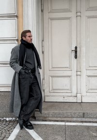 Szary płaszcz, ciemny golf, czarne dżinsy i buty. Mężczyzna opiera się o drzwi z teksturowanego drewna, otoczony kamiennymi stopniami i chodnikiem.