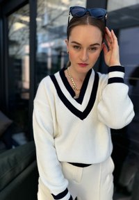 Λευκό πλεκτό πουλόβερ με navy V-λαιμόκοψη και ρίγες. Συνδυάζεται με ανοιχτόχρωμα παντελόνια και χρυσή αλυσίδα στο λαιμό.