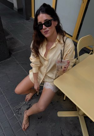 Mujer con gafas de sol y una camisa amarillo pálido está sentada al aire libre sosteniendo café con hielo cerca de una mesa y silla amarillas.