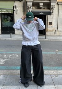 Grå hettejakke med grønne grafiske elementer, hvit lagdelt skjorte og sorte bukser med vide ben. Grønn caps og svarte sko med hvite striper. Gateomgivelse.