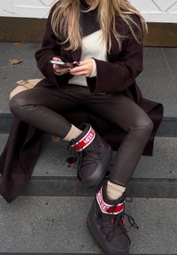 Άτομο με μακριά ξανθά μαλλιά που φοράει σκούρο παλτό, λευκό πουλόβερ, δερμάτινο παντελόνι και μαύρα Moon Boots, καθισμένο σε γκρι πέτρινα σκαλιά, χρησιμοποιώντας ένα smartphone.