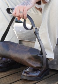 Main tatouée tenant une sangle de sac en cuir noir, personne portant un pantalon beige et des chaussures en cuir brun foncé assise sur des marches en pierre.