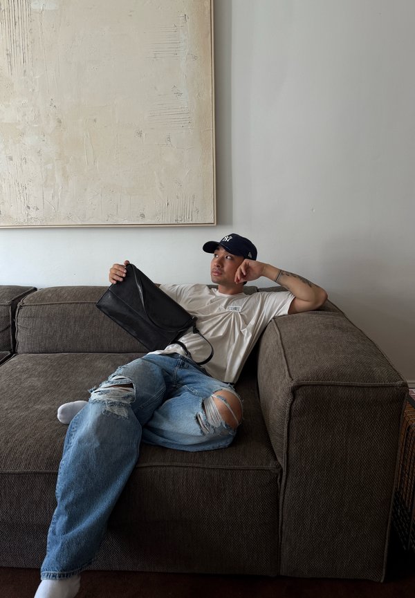 Jeune homme portant une casquette et un jean déchiré, allongé sur un canapé marron, tenant un sac noir avec un grand tableau beige texturé derrière lui.