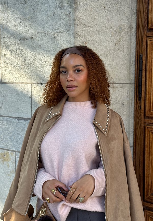 Giacca di suede marrone con borchie argentate, maglione rosa e pantaloni grigi. Capelli ricci, indossa anelli e tiene degli occhiali da sole. Dettagli del tessuto testurizzato visibili.