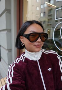 Vinrød kordfløyelsjakke med glidelås, hvite striper, store svarte solbriller med brune linser, og sølvstore øredobber.