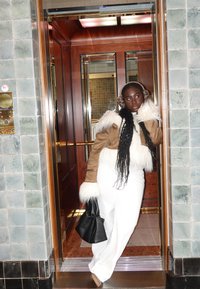 Veste en shearling marron avec bordure en fourrure blanche, pantalon blanc à jambes larges, et sac à main noir. Debout dans un ascenseur au intérieur en bois et carrelage.