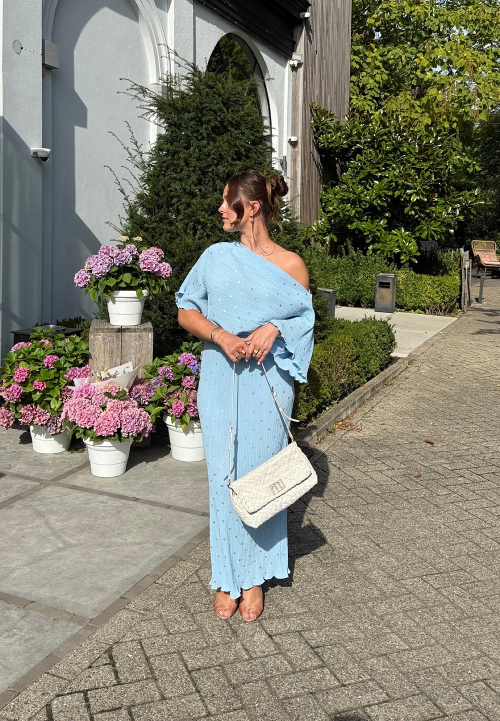 Vestido azul claro com design off-shoulder, tecido texturizado e pérolas decorativas. Bolsa branca com alça de corrente. Fundo floral.