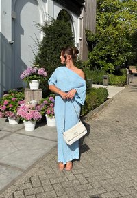 Vestido azul claro com design off-shoulder, tecido texturizado e pérolas decorativas. Bolsa branca com alça de corrente. Fundo floral.