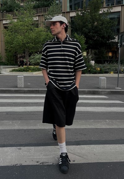 Polo a rayas blanco y negro con media cremallera, pantalones cortos negros y zapatos Adidas negros, de pie en un paso de peatones.