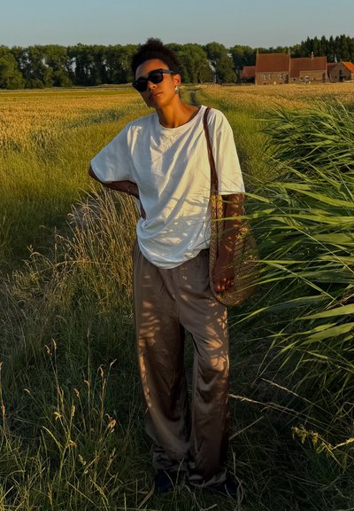 Camiseta blanca de algodón suelta, pantalones anchos de satén beige, gafas de sol negras y una bolsa tejida, de pie en hierba alta con un fondo rural.