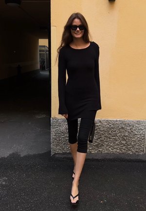 Vestido negro de manga larga con canalé, combinado con leggins negros cortos y chanclas negras. La modelo sostiene un bolso de mano y está de pie frente a una pared amarilla.