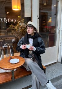 Veste en cuir noire oversize, béret blanc, écharpe noire, pantalon à carreaux, tenant une tasse de café. Table en bois avec une petite assiette blanche et un briquet.