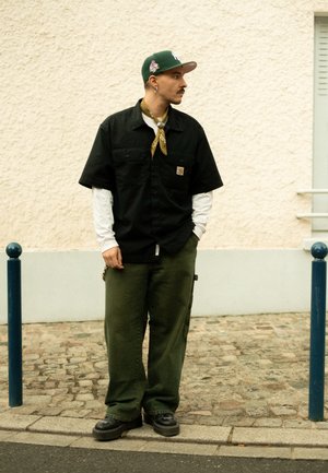 Svart kortärmad skjorta, gröna byxor, gul bandana och en grön keps. Svarta skor med tjock sula. Står bredvid en blå stolpe.