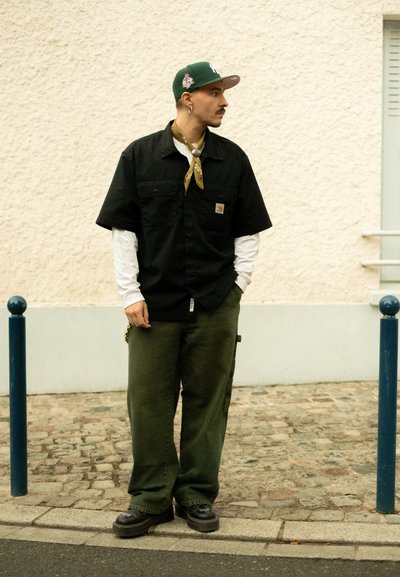 Camisa negra de manga corta, pantalones verdes, bandana amarilla y gorra verde. Zapatos negros con suela gruesa. De pie junto a un poste azul.