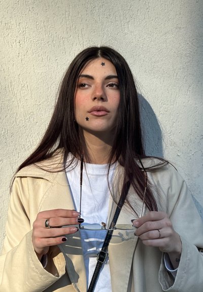 Mujer joven con cabello largo y oscuro que lleva un abrigo beige y una camisa blanca, sosteniendo gafas, con dos pegatinas en forma de estrella en la cara, frente a una pared texturizada.