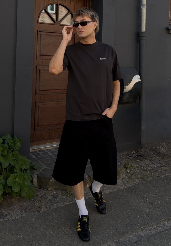 Svart kortermet t-skjorte med logo, løse svarte shorts, hvite sokker og svarte sneakers med gule striper, mot en trevegg.