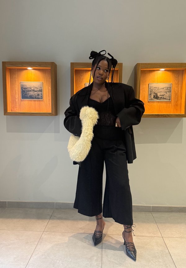 Jeune femme aux cheveux coiffés porte un blazer noir oversize, un pantalon à jambes larges, des escarpins lacés et porte un sac crème duveteux, debout près de photos encadrées éclairées.