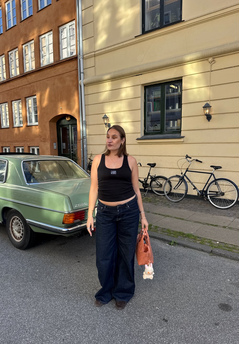 Černá zkrácená tílko, široké tmavé džíny, hnědé boty. Oranžová taška s plyšovým hračkou. Zelené vintage auto v pozadí poblíž žluté budovy.