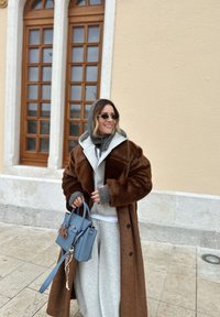 Femme portant un manteau en fourrure marron et un sweat à capuche gris, tenant un sac à main bleu, debout près d'un bâtiment beige avec de grandes fenêtres en bois.