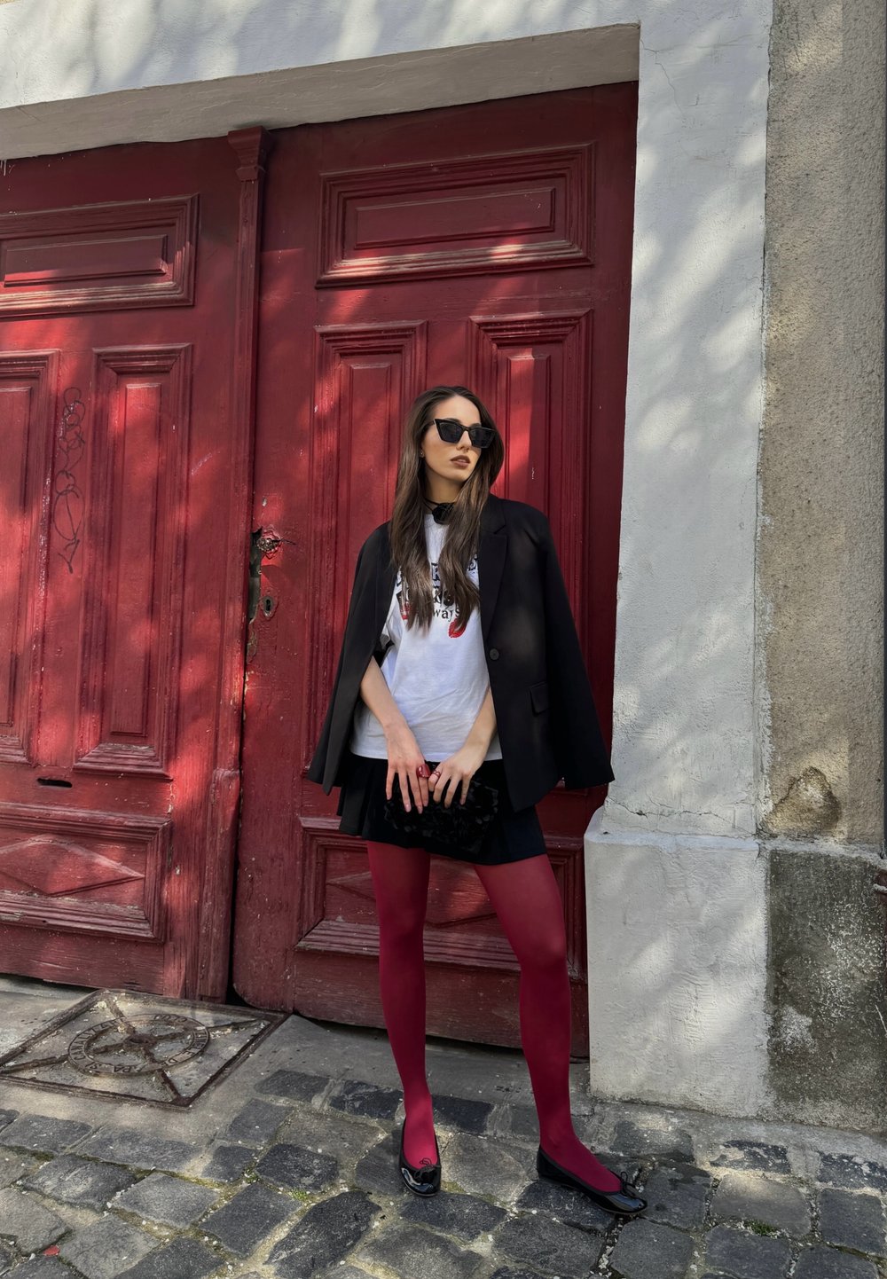 Czarna marynarka na białej koszulce z grafiką, czarna spódnica i bordowe rajstopy. Zestaw dopełniają czarne płaskie buty i teksturowana czarna kopertówka. W tle czerwone drzwi.